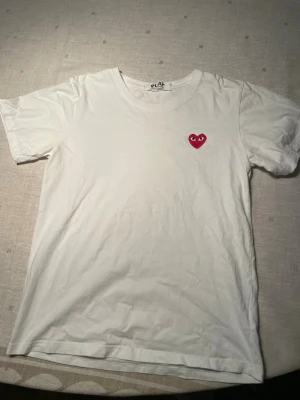 Vit t-shirt från Comme des Garçons Play - Vit t-shirt från Comme des Garçons Play med det ikoniska röda hjärtat med ögon broderat på bröstet. Klassisk passform och rund halsringning. Perfekt för dig som gillar stilrena plagg med en twist. Storlek S 