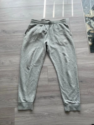 Gråa mjukisbyxor från Nike - Snygga grå mjukisbyxor från Nike med klassisk logga broderad på vänster ben. Byxorna har resår i midjan, dragsko och muddar vid bensluten. Tillverkade i mjukt och bekvämt material, perfekta för en avslappnad stil.