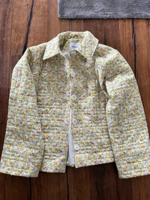 Blommig quiltad jacka från Envii - Quiltad jacka från Envii med blommigt mönster i ljusa toner av gul, grön och lila. Jackan har stora vita knappar framtill, klassisk krage och lång ärm. Perfekt för dig som gillar en söt och trendig stil.