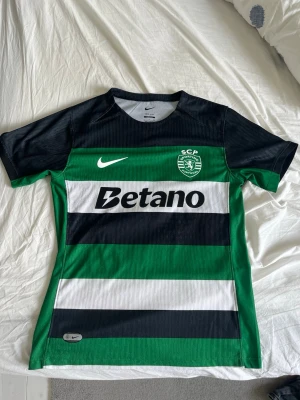 Sporting CP matchtröja Nike Gyökeres - Säljer en officiell Sporting CP matchtröja från Nike med Gyökeres och nummer 9 på ryggen. Tröjan har gröna, svarta och vita breda ränder, korta ärmar och klubbmärke på bröstet. Tillverkad i lätt och ventilerande material, perfekt för fotbollsträning eller match.