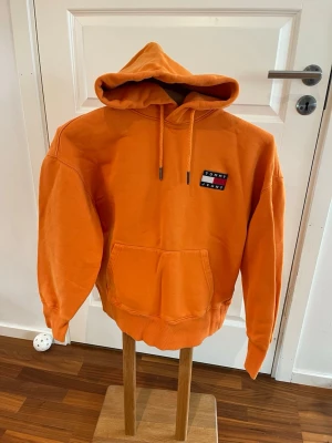 Orange hoodie från Tommy Jeans - Snygg orange hoodie från Tommy Jeans med klassisk känguruficka och justerbar huva med dragsko. Liten broderad logga på bröstet i rött, vitt och blått. Tillverkad i mjuk bomullsblandning och har ribbade muddar vid ärmar och midja.