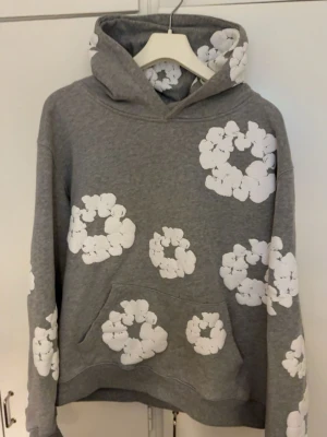 Grå hoodie från denim tears  - Grå hoodie med stora vita blommönster över hela tröjan, inklusive huvan. Har en klassisk känguruficka framtill och ribbade muddar vid ärmslut och nederkant. Perfekt för en avslappnad och trendig stil.