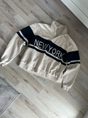 Beige sweatshirt med New York-tryck - Snygg beige sweatshirt med mörkblåa ränder och stort 'NEW YORK'-tryck över bröstet. Tröjan har hög krage utan dragkedja, långa ärmar och muddar vid ärmslut och nederkant. Perfekt för en avslappnad streetwear-look.