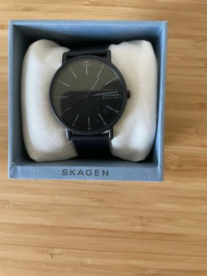 Svart minimalistisk klocka från Skagen - Stilren svart klocka från Skagen med rund urtavla och diskreta vita markeringar. Klockan har ett svart läderband och enkel, modern design som passar till det mesta. Perfekt för dig som gillar minimalistisk stil.