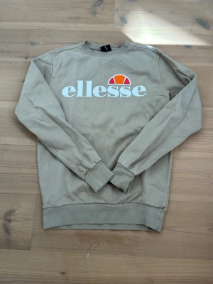 Beige sweatshirt från Ellesse - Snygg beige sweatshirt från Ellesse med stor logga framtill i vitt och orange. Tröjan har rund hals, långa ärmar och ribbade muddar vid ärmslut och nederkant. Perfekt för en avslappnad och sportig stil.