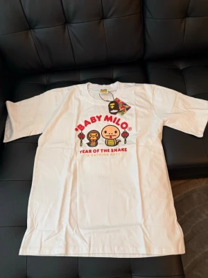 Vit Baby Milo t-shirt från BAPE - Vit t-shirt från A Bathing Ape med Baby Milo-tryck och texten 'Year of the Snake' framtill. Klassisk passform med rund hals och korta ärmar. Materialet är mjuk bomull och trycket har färgglada detaljer.