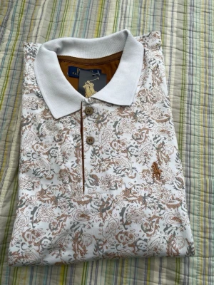 Vit mönstrad pikétröja Polo Ralph Lauren - Vit pikétröja från Polo Ralph Lauren med brunt och grått blommönster, klassisk krage och två knappar framtill. Broderad logga på bröstet. Perfekt för en stilren och avslappnad look.