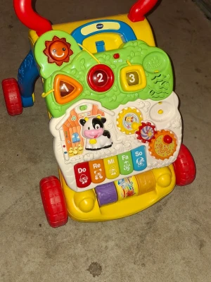 Färgglad gåvagn med leksakspanel - Rolig och färgglad gåvagn från VTech med interaktiv leksakspanel. Panelen har knappar, snurrhjul, former, siffror och musikknappar i olika färger. Vagnen har röda hjul, blåa och gula detaljer samt ett rött handtag. Perfekt för små barn som lär sig gå.