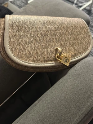 Beige Michael Kors handväska - Snygg beige handväska från Michael Kors med MK-mönster över hela väskan. Väskan har rundade kanter, guldfärgad detalj med MK-logga och är tillverkad i canvas med läderdetaljer. Perfekt accessoar för att lyfta din outfit.