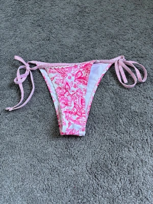 Bikini underdel - Söt bikiniunderdel i rosa och vitt med blommigt mönster och knyt i sidorna. Perfekt för sommaren och strandhäng. Tunn och lätt design som ger en bekväm passform.