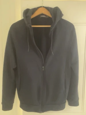 Mörkblå zip-hoodie med huva - En stilren mörkblå hoodie med dragkedja från Dressmann. Hoodien har huva med dragsko, långa ärmar och ribbade muddar. Perfekt för en avslappnad look och enkel att matcha med jeans eller joggers. Använd 2-4 gånger, hängd i garderoben resten utav tiden! :)