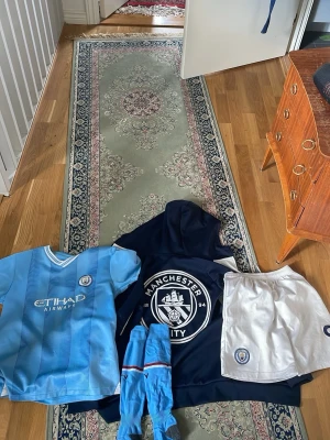 Manchester City matchkit och hoodie - Säljer ett komplett Manchester City matchkit med ljusblå matchtröja, vita shorts, ljusblå strumpor och en marinblå hoodie med klubbens logga. Ingår även en zip-hoodie i vitt och marinblått med klubbmärke. Perfekt för fotbollsträning eller support.