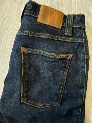 Nudie Jeans  - Säljer ett par mörkblå jeans från Nudie Jeans med klassisk femficksmodell och kontrastsömmar i orange. Jeansen har rak passform och är tillverkade i kraftig denim. Snygg läderpatch med logga bak i midjan. Perfekta till en avslappnad streetwear-look.