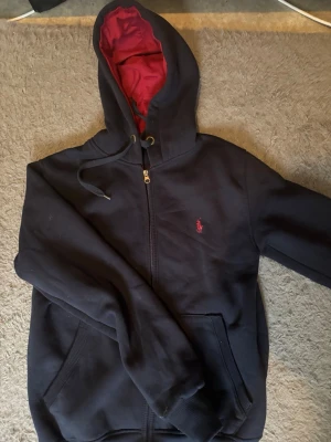 Mörkblå hoodie från Polo Ralph Lauren - Mörkblå hoodie med dragkedja från Polo Ralph Lauren. Har röd insida i huvan, dragsko och klassisk broderad logga på bröstet. Mjuk bomullskänsla och två fickor framtill. Perfekt för en chill och stilren look.