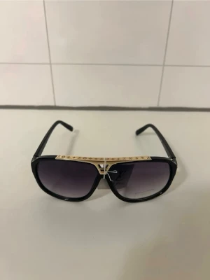 Svarta Louis Vuitton solglasögon - Snygga svarta solglasögon från Louis Vuitton med guldiga detaljer och en unik metallbåge över näsan. Glasen är mörktonade och bågarna har en klassisk, fyrkantig form. Perfekt accessoar för att lyfta din stil.