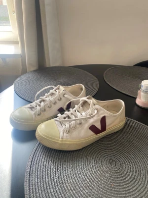 Vita Veja sneakers med vinrött V - Snygga vita sneakers från Veja med klassisk låg profil och vinrött V på sidan. Skorna har vita snören, gummisula och tåhätta samt ovandel i canvas. Perfekta för dig som gillar stilrena och trendiga sneakers med en clean look.