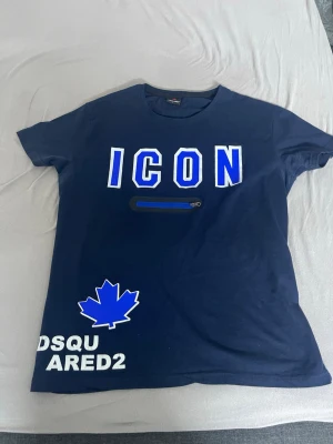 Blå Dsquared2 ICON t-shirt - Mörkblå t-shirt från Dsquared2 med stor vit och blå 'ICON'-text på bröstet, en liten dragkedjedetalj och ett blått lönnlöv. Tryckt logga nere till vänster. Klassisk passform och rund hals. Perfekt för dig som gillar streetwear.