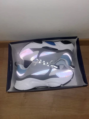 Dior B22 sneakers grå/blå/vit - Chunky Dior B22 sneakers i grått, vitt och blått med mesh och läderdetaljer. Snygg design med tjock sula och tydlig Dior-logga på sidan. Perfekta för dig som gillar streetwear och exklusiva märken.