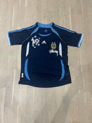 Argentina Adidas fotbollströja S - Mörkblå Argentina fotbollströja från Adidas med ljusblå och vita detaljer, nummer 19 och AFA-emblem på bröstet. Tröjan har Stüssy-logga och är tillverkad i lätt, ventilerande material. Perfekt för fotboll eller sportiga outfits.
