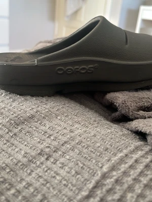 Svarta tofflor från Oofos - Svarta tofflor från Oofos med öppen tå och bred rem över foten. Tillverkade i ett mjukt och stötdämpande syntetmaterial som ger skön komfort. Perfekta att glida i hemma eller efter träning. Snygg minimalistisk design med Oofos-logga i sulan.