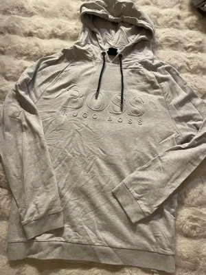 Ljusgrå hoodie från Hugo Boss - Snygg ljusgrå hoodie från Hugo Boss med broderad logga på bröstet. Hoodien har dragsko i huvan och ribbade muddar vid ärmslut och nederkant. Perfekt för en avslappnad och stilren look.
