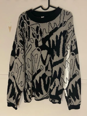 Svart och grå mönstrad stickad tröja - Cool stickad tröja med abstrakt svart och grått mönster. Oversized passform med rund halsringning och ribbade muddar. Perfekt för dig som gillar streetstyle och unika plagg.