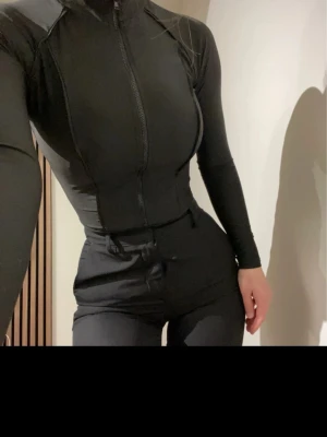 Svart body med dragkedja - Säljer  svart body med långärmad topp och högmidjade tights. Bodyn har dragkedja framtill och stårkrage, materialet är stretchigt och följsamt. Perfekt för träning eller gym. Snygg och tight passform.