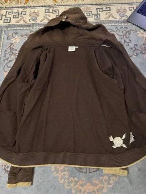 Brun hoodie med tryck och huva - Snygg brun hoodie från WESC med huva och ribbade muddar i beige. Har coola tryckdetaljer i beige på både fram- och baksidan samt på ena ärmen. Perfekt för dig som gillar streetwear och vill ha en unik look.