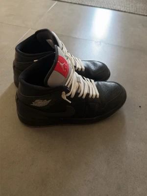 Svarta Air Jordan 1 High sneakers - Snygga svarta Air Jordan 1 High sneakers med vita skosnören och röd-vit tunga. Klassisk vinge-logga på sidan och svart swoosh. Skorna har en hög siluett och är tillverkade i läder för en clean och sportig look som är helt nya