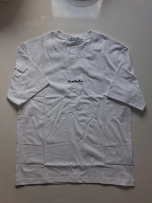 Vit Acne Studios t-shirt i bomull - Vit t-shirt från Acne Studios med minimalistisk svart logga broderad på bröstet. Klassisk passform med rund hals och korta ärmar. Tillverkad i mjuk bomull, perfekt för en clean och stilren look.