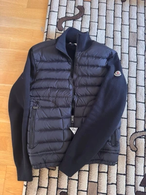 Svart cardigan jacka från Moncler - Snygg svart pufferjacka från Moncler med stickade ärmar och vadderad front. Jackan har dragkedja framtill, två fickor med dragkedja och Moncler-logga på ärmen. Perfekt för kalla vinterdagar och ger en stilren look.