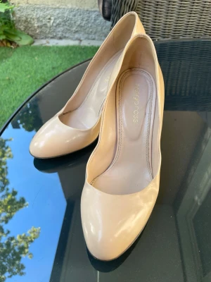 Beige pumps från Sergio Rossi - Snygga pumps i beige lack från Sergio Rossi. Klassisk rund tå och hög klack ger en elegant siluett. Skorna är tillverkade i glansigt material som ger en lyxig känsla och passar perfekt till många olika outfits.
