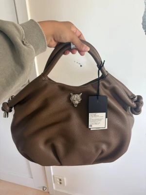 Brun handväska från Plein Sport - Snygg brun handväska i mjukt, strukturerat konstläder från Plein Sport. Väskan har rundade handtag, knutdetaljer på sidorna och ett coolt silverfärgat varghuvud framtill. Perfekt för dig som vill ha en trendig och rymlig väska med unika detaljer.