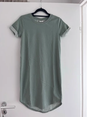 Ljusgrön t-shirtklänning från JDY - En enkel och stilren ljusgrön t-shirtklänning från JDY. Klänningen har rund halsringning, korta ärmar med uppvik och rak passform. Perfekt för en avslappnad look under varmare dagar.