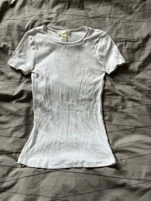 Vit ribbad t-shirt från H&M - En klassisk vit ribbad t-shirt från H&M i storlek XS. T-shirten har korta ärmar, rund halsringning och är tillverkad i mjuk bomull. Perfekt basplagg att matcha med allt och har en slim passform som sitter snyggt 😊🩷
