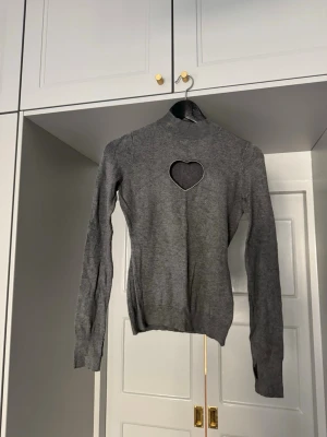 CLOEYS hjärtformad cut-out - Trendig grå stickad tröja med lång ärm och hög hals. Framtill finns en hjärtformad cut-out som ger en unik och snygg detalj. Perfekt för dig som vill ha något extra i garderoben. Materialet är mjukt och följsamt.