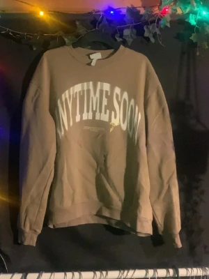 Brun sweatshirt med tryck - Cool brun sweatshirt med stort vitt tryck på bröstet där det står 'ANYTIME SOON'. Tröjan har en avslappnad passform och ribbade muddar vid ärmslut och nederkant. Perfekt för en chill och trendig look.