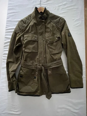 Belstaff Gold Label Made in Italy vaxad jacka - Exklusiv vintagejacka från Belstaff Gold Label, tillverkad i Italien. Klassisk field jacket-modell med bälte i en tidlös khakifärg, utility-detaljer och rutigt innerfoder. Tillverkad i kraftigt kvalitetsbomullstyg med slitstark känsla som håller i många år.  Jackan har flera framfickor, justerbart midjebälte, tryckknappar och klassiska Belstaff-detaljer. Perfekt för vardagsstil, biker-look eller klassisk vintage outfit.  Tillverkad i Italien.  Fint vintage skick med naturliga bruksspår som ger j