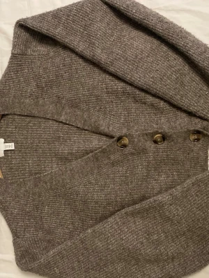 Gråmelerad kofta från H&M - Mysig gråmelerad kofta från H&M med v-ringning och stora bruna knappar framtill. Koftan är stickad i mjukt material och har långa ärmar samt ribbade muddar vid ärmslut och nederkant. Perfekt för lager på lager och chill dagar.Koftan är även kort i modellen 