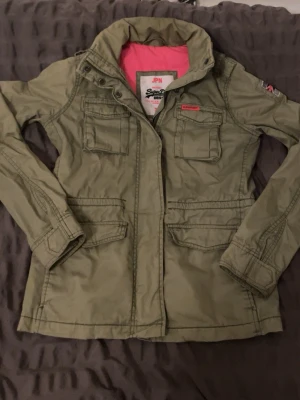 Olivgrön utilityjacka Superdry - Säljer en olivgrön utilityjacka från Superdry med fyra fickor framtill, dragkedja och tryckknappar. Jackan har en snygg rosa innerfoder och patchdetaljer på ärmen. Perfekt för dig som gillar streetwear och funktionell stil.