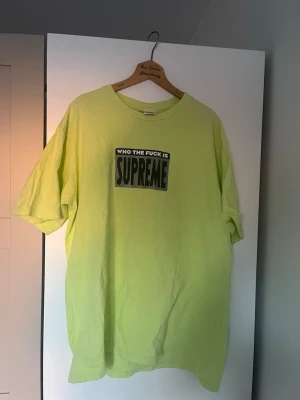 Gul/neongrön Supreme t-shirt med tryck - Säljer en ljusgul t-shirt från Supreme med stort svart och vitt tryck på bröstet där det står 'WHO THE FUCK IS SUPREME'. T-shirten har klassisk passform, rund hals och är tillverkad i mjuk bomull. Perfekt för dig som gillar streetwear och statement-plagg. Storlek L