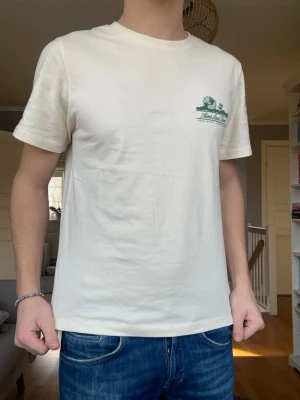 Aimé Leon Doré T-shirt – strl S - Snygg T-shirt från Aimé Leon Doré med diskret bröstprint och stor graphic på ryggen. Väldigt clean och eftertraktad modell från ett av de hetaste streetwear-märkena just nu.  Bra passform, funkar perfekt till jeans, chinos eller shorts.  Öppen för bud! Skick: Mycket bra. Storlek: S. Färg: Off-white / vit med grön print. Pris: 399 kr 