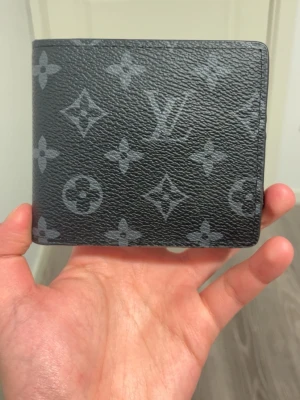 L’’v svart monogram plånbok - Snygg svart plånbok från L’’v i klassiskt monogrammönster. Tillverkad i tåligt läder med gråa L’’Vloggor och blommor över hela ytan. Perfekt storlek för fickan och stilren design som sticker ut.