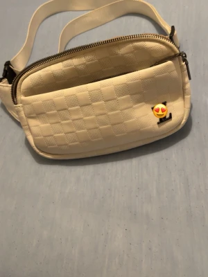 Vit axelväska med rutmönster - Snygg vit axelväska i konstläder med rutmönstrad struktur och två dragkedjefack. Väskan har en emoji-pin framtill och justerbar axelrem. Perfekt för att bära det viktigaste med stil.