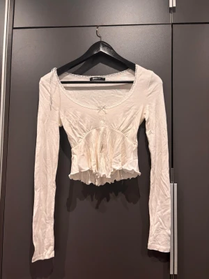 Vit långärmad topp Gina Tricot - Söt vit topp från Gina Tricot med lång ärm och volangkant nedtill. Toppen har en liten rosettdetalj framtill och en djup rundad urringning med skuren slits vid magen. Aldrig använd men lite skrynklig pga legat länge. 