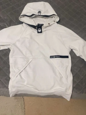 Vit hoodie från Sail Racing - Snygg vit hoodie från Sail Racing med stor huva och svarta detaljer. Hoodien har en praktisk magficka och en logotyp på bröstet. Perfekt för dig som gillar en clean och sportig stil. Materialet känns mjukt och bekvämt.