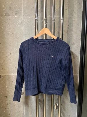 Mörkblå kabelstickad tröja Gant - Mörkblå kabelstickad tröja från Gant med rund halsringning och diskret logga på bröstet. Tröjan har långa ärmar och ribbade muddar vid ärmslut och nederkant. Perfekt för en klassisk och stilren look. Den är passar dig som är 10-12 år den är storlek 142-152