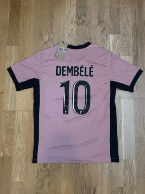 PSG Dembélé matchtröja rosa/svart - Snygg PSG matchtröja i rosa med svarta detaljer och tryck. Dembélé och nummer 10 på ryggen. Framsidan har PSG-logga, Jordan-logga och Qatar Airways sponsortryck. Tillverkad i lätt och ventilerande material, perfekt för fotboll eller träning.