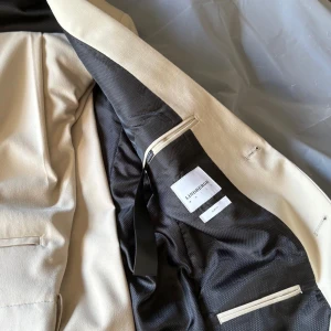 Beige kavaj från Lindbergh, storlek 50 - Snygg beige kavaj från Lindbergh med klassisk passform och två svarta knappar framtill. Kavajen har fickor med lock, slits bak och svarta knappar vid ärmslut. Insidan är fodrad i svart och har innerficka med dragkedja. Perfekt för en stilren look.