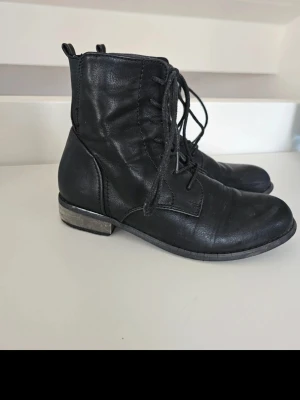 Svarta snörade boots - Säljer ett par svarta stövletter med snörning framtill och dragflikar bak. Skorna har låg klack och är gjorda i ett matt, skinnliknande material. Mindre Skavanker i klacken. Fler bilder kan skickas vid intresse!